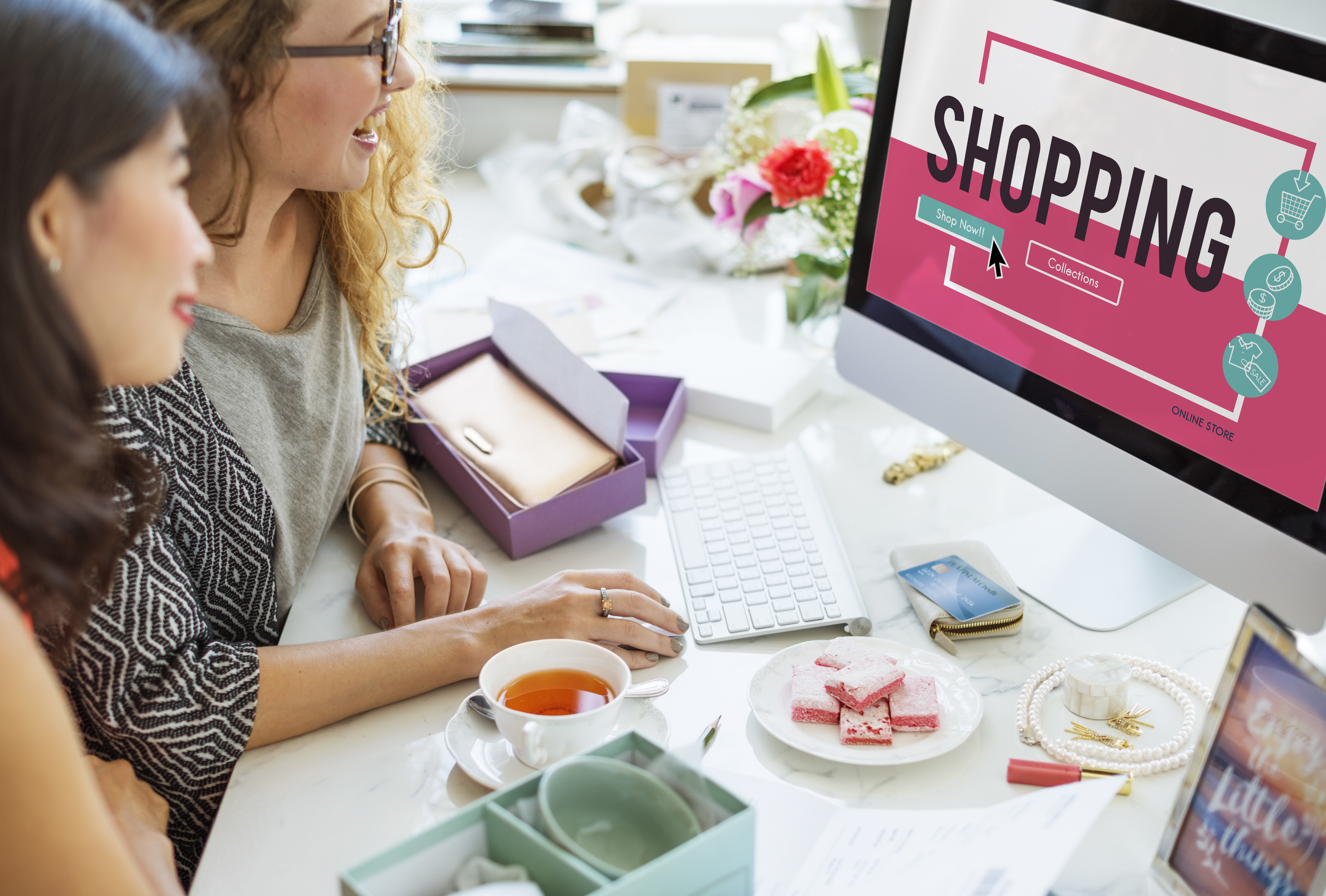 Two women shopping online on desktop computer browsing eCommerce store with credit card and products on desk.